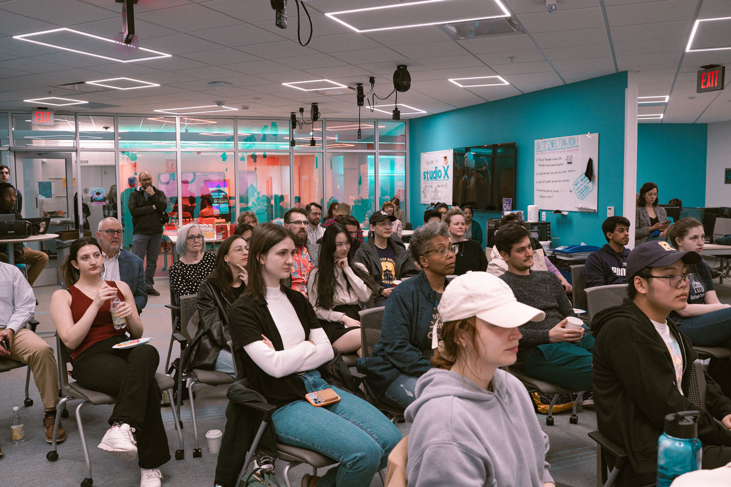 A large audience sitting in chairs in Studio X listens to presentations.