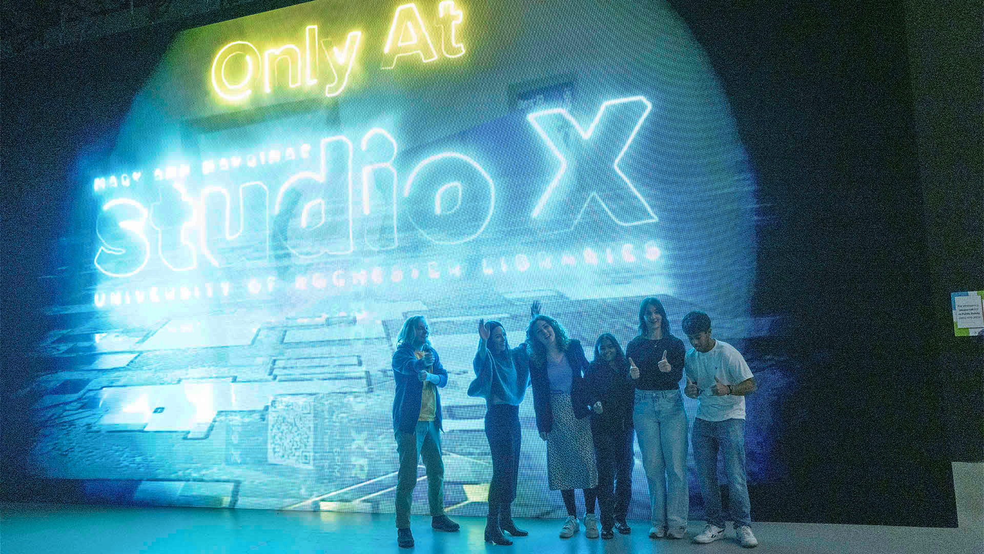 Group of students posing in front of massive digital board reading Studio X.